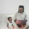 A father working on a laptop at home as his toddler daughter sits beside him, highlighting family balance.