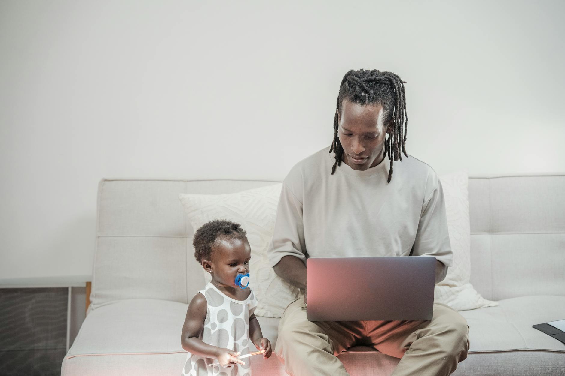 A father working on a laptop at home as his toddler daughter sits beside him, highlighting family balance.