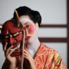 A geisha in a kimono holding a Hannya mask, symbolizing traditional Japanese culture.