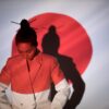 A stylish woman in a white blazer stands before a Japanese flag silhouette, looking down.