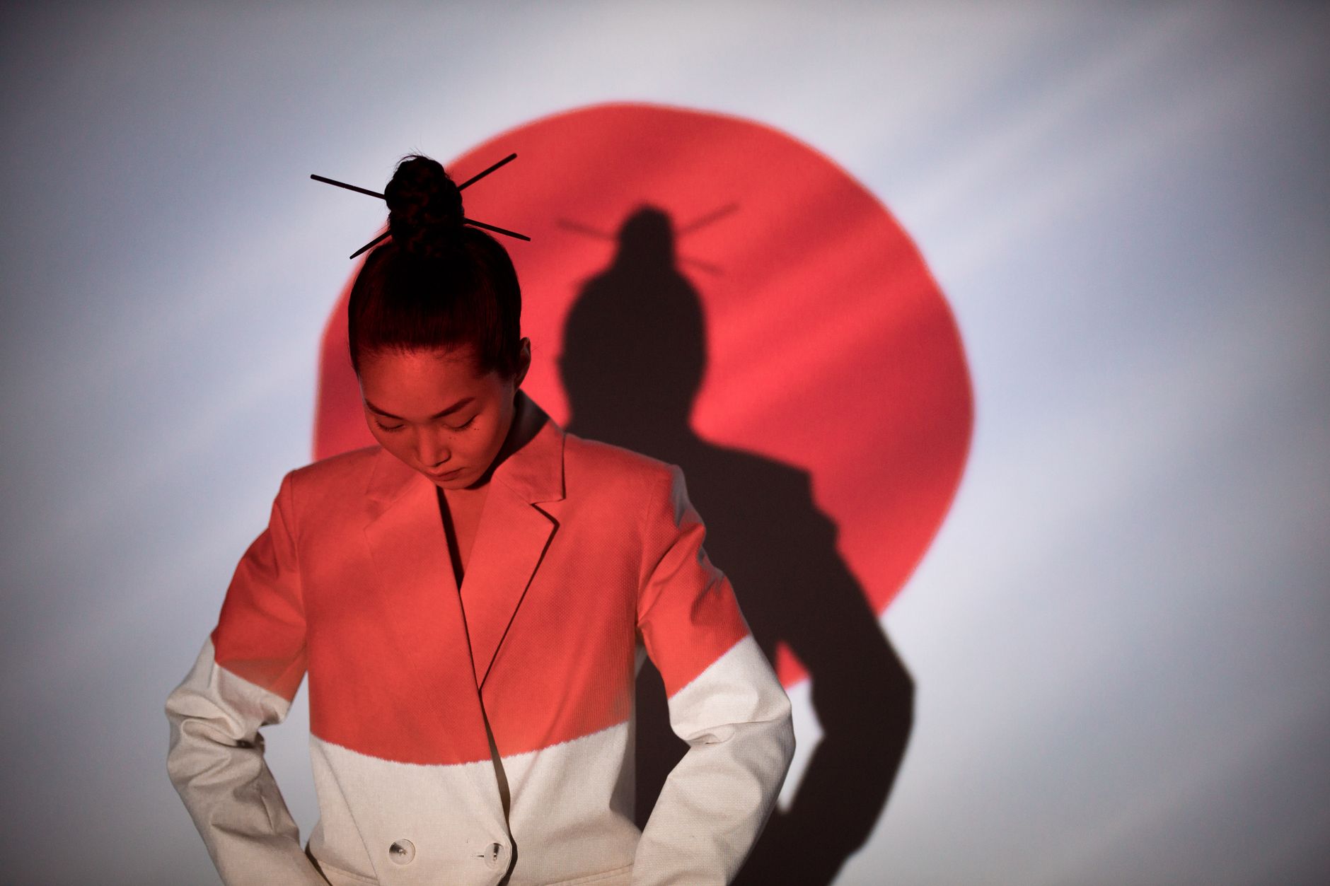 A stylish woman in a white blazer stands before a Japanese flag silhouette, looking down.