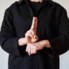 A person in a black coat demonstrating a sign language gesture indoors.