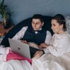 A bride and groom sitting indoors, browsing a laptop together in wedding attire.