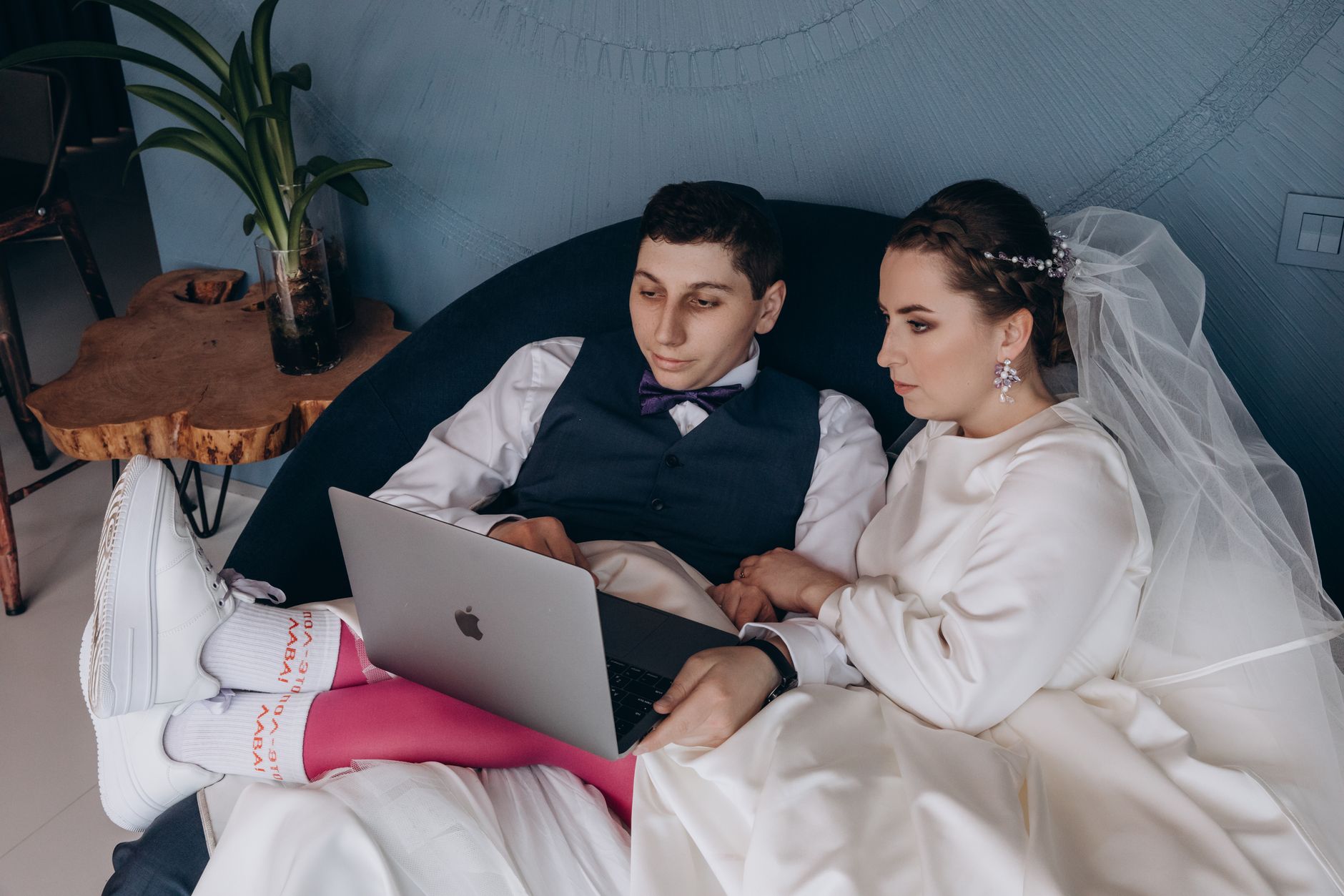 A bride and groom sitting indoors, browsing a laptop together in wedding attire.