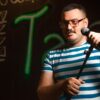 Man holding a microphone doing stand-up comedy in casual striped shirt on stage.