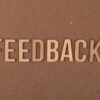 A close-up of a wooden 'FEEDBACK' sign on a textured cardboard background.