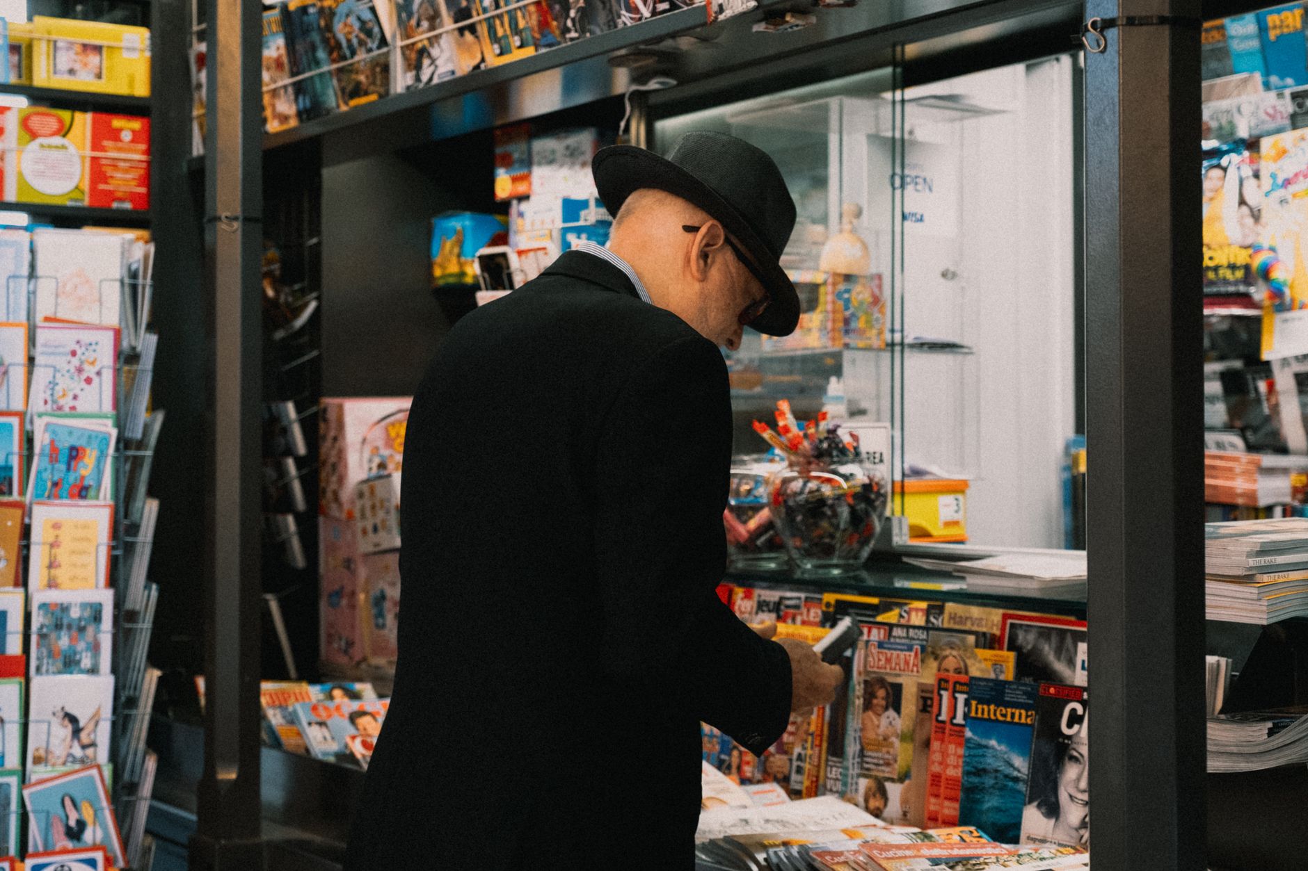 A senior man in a hat browsing magazines at a kiosk. Indoor setting with diverse print media.