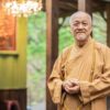 Portrait of an elderly Buddhist monk wearing a traditional robe, standing calmly in a serene setting.