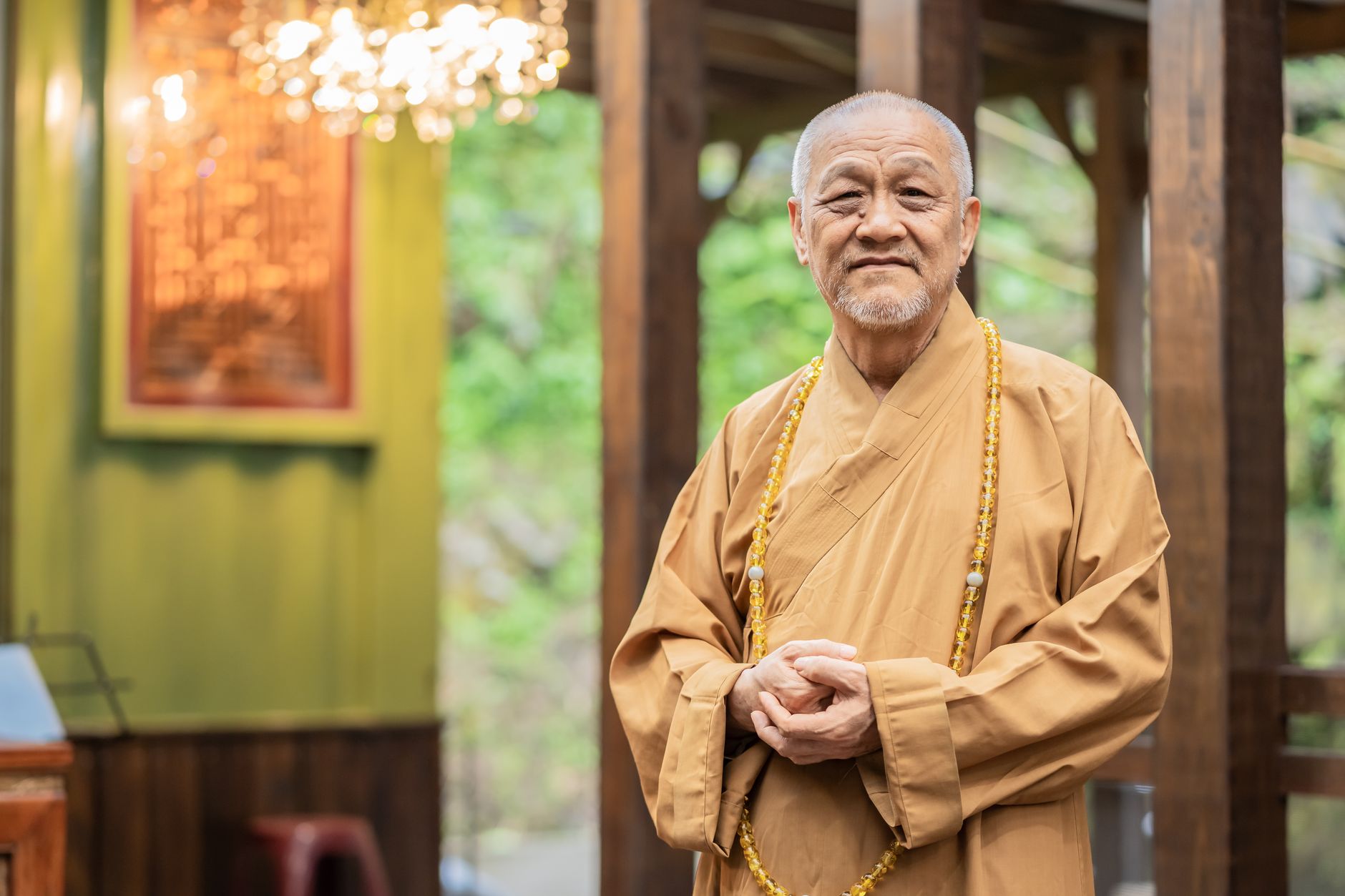 Portrait of an elderly Buddhist monk wearing a traditional robe, standing calmly in a serene setting.