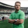 Man in green polo stands confidently with arms crossed in a sports stadium.