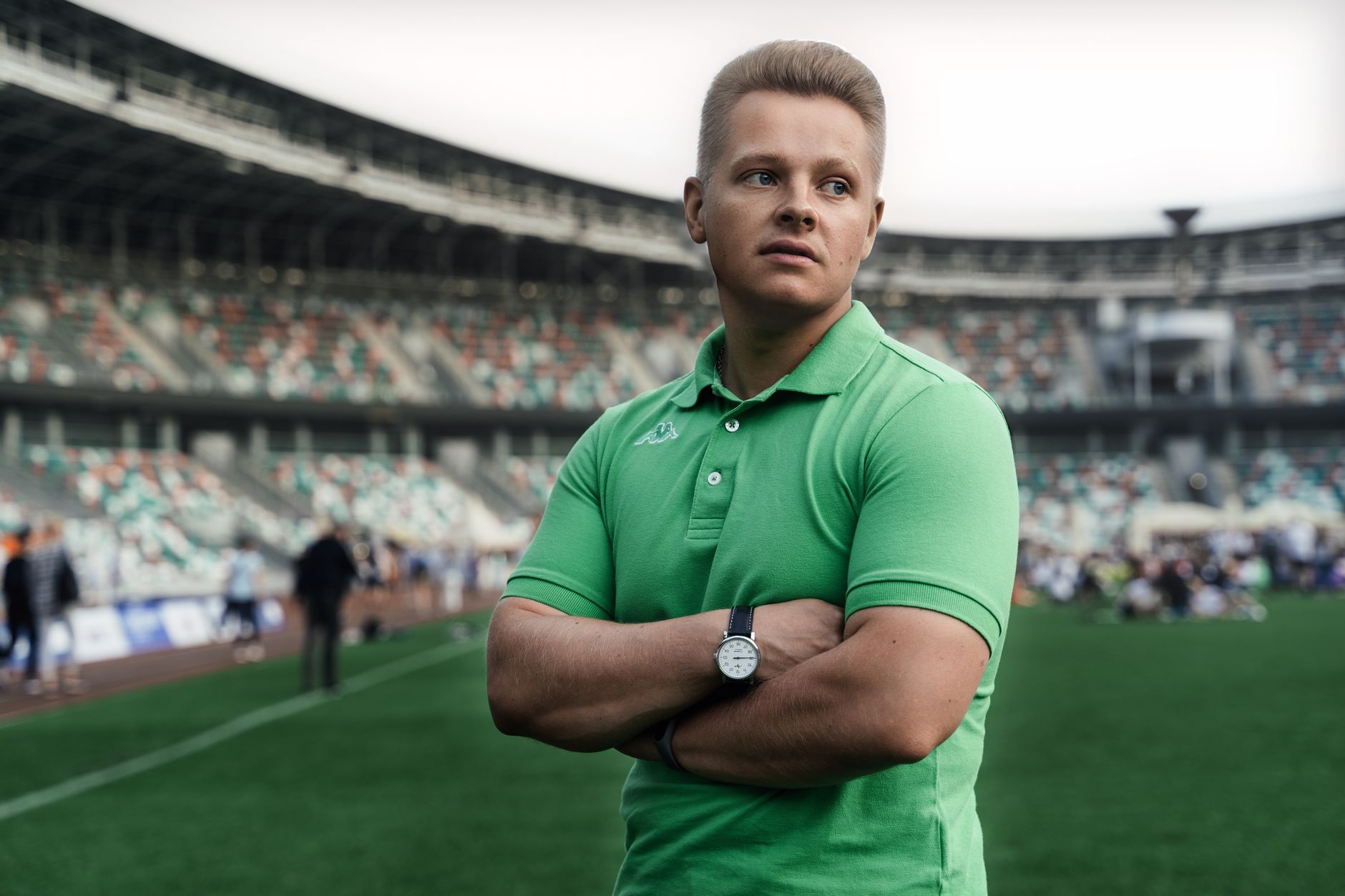 Man in green polo stands confidently with arms crossed in a sports stadium.