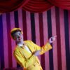 Cheerful clown in a vibrant yellow suit pointing during a stage performance, against a striped background.