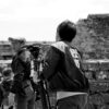 Black and white image of a cameraman filming near stone walls outdoors, with people in the background.