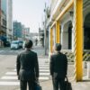 Two businessmen in suits crossing a street in an urban Japanese setting, reflecting daily city life.