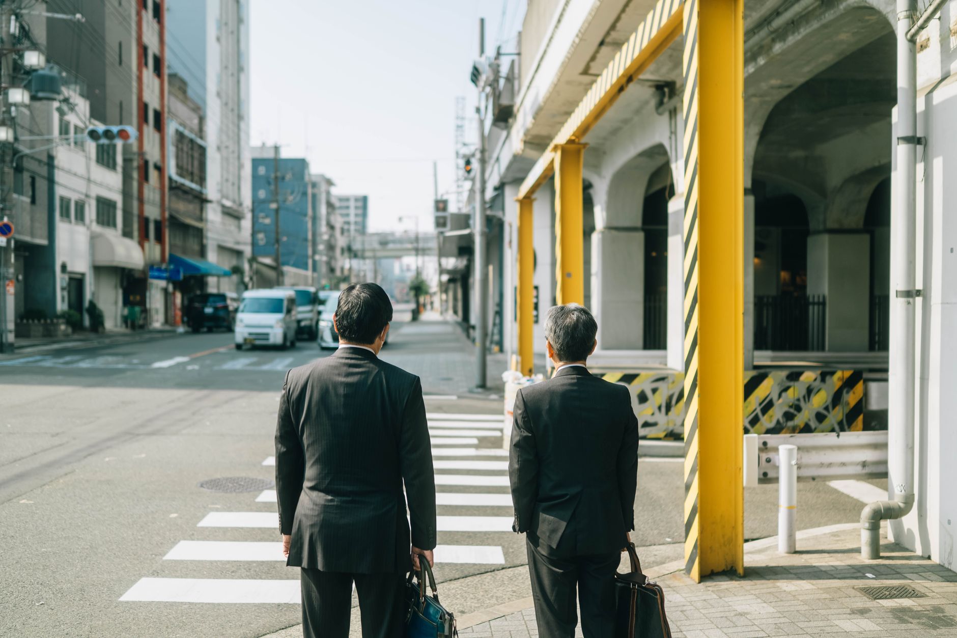 Two businessmen in suits crossing a street in an urban Japanese setting, reflecting daily city life.