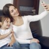 Delighted mother and daughter in casual wear sitting on sofa and taking selfie while resting in contemporary living room during daytime