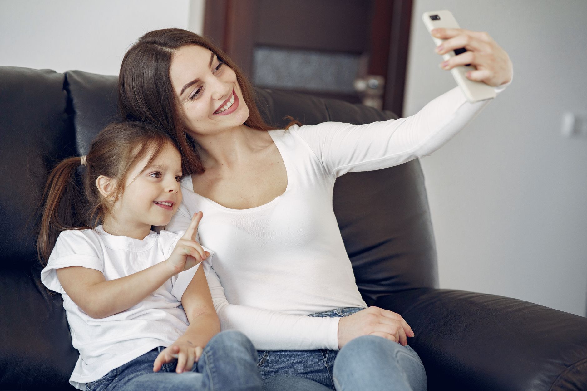 Delighted mother and daughter in casual wear sitting on sofa and taking selfie while resting in contemporary living room during daytime