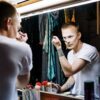 A man applies makeup in a dressing room mirror, showcasing transformation and creativity.