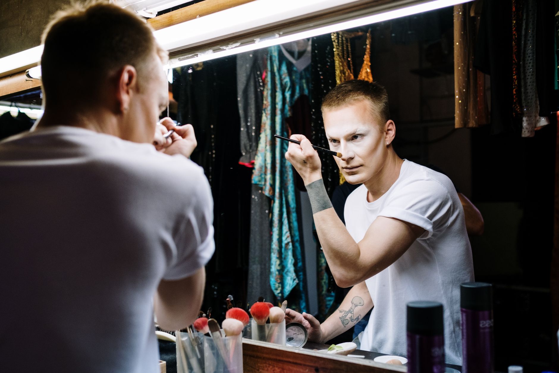 A man applies makeup in a dressing room mirror, showcasing transformation and creativity.