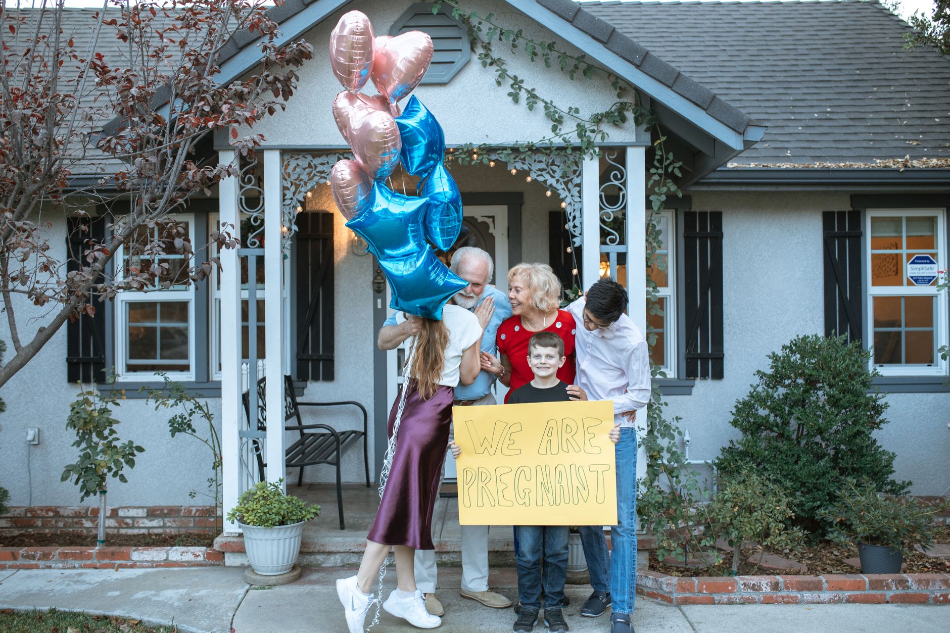Family joyfully announcing pregnancy with balloons in front of house.