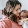 Side view of caring mother in casual clothes with adorable infant in arms sitting on comfortable sofa in light room at home