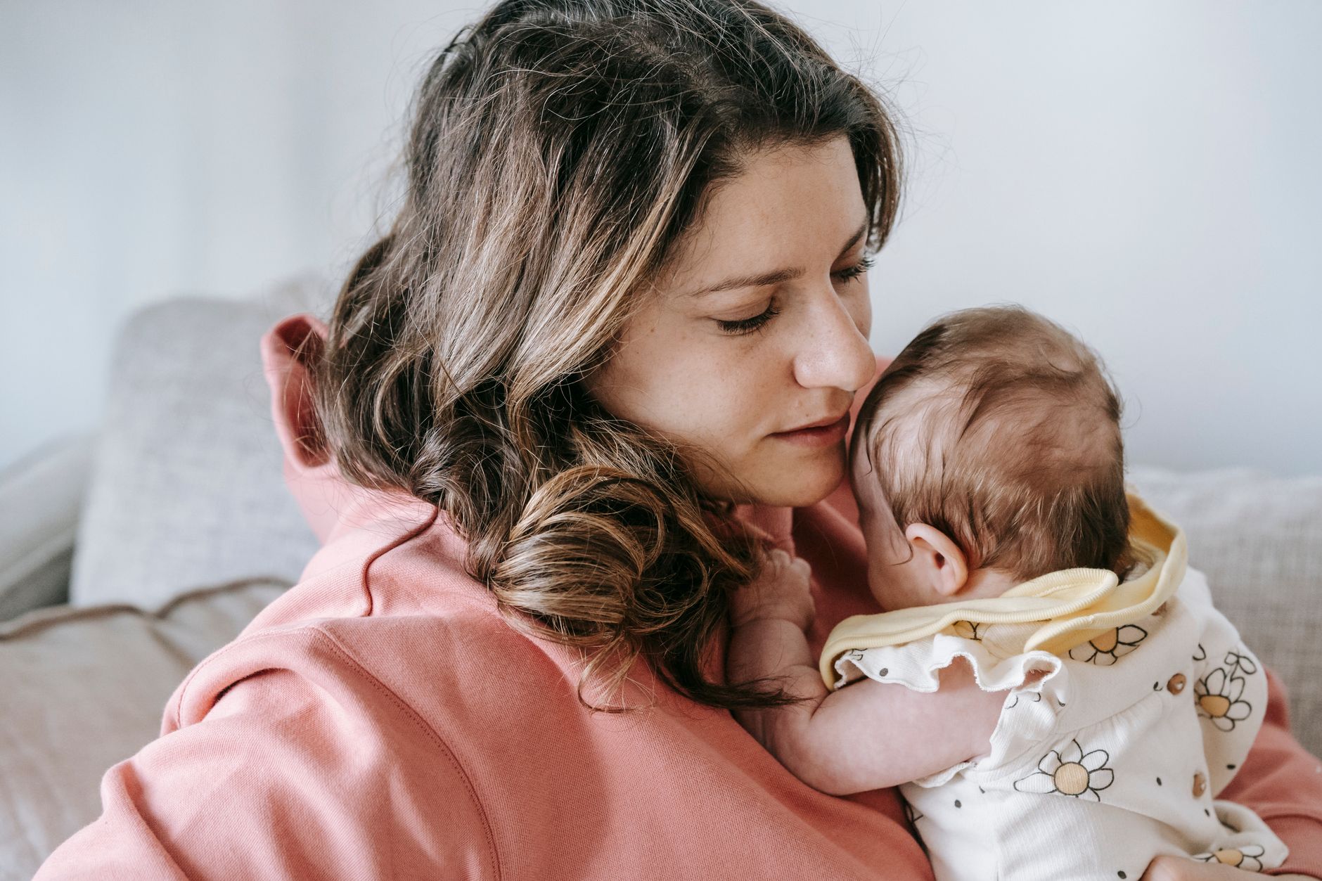 Side view of caring mother in casual clothes with adorable infant in arms sitting on comfortable sofa in light room at home