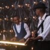 Three musicians perform with piano, guitar, and drums in a stylish decorated room.