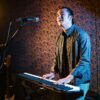 A male musician playing keyboard piano on stage under studio lights indoors.