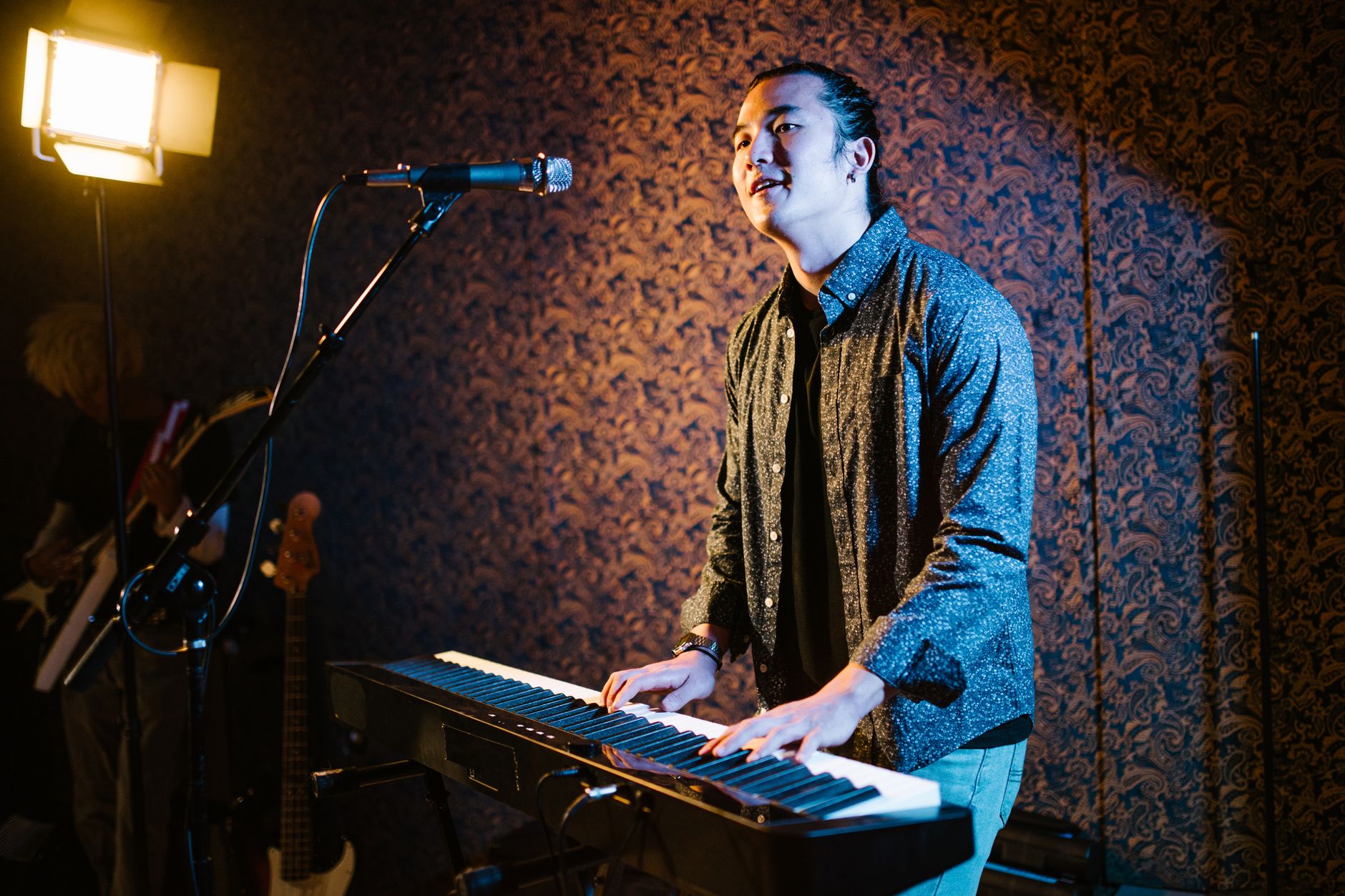 A male musician playing keyboard piano on stage under studio lights indoors.