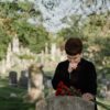 A woman in black paying her respects at a grave, holding flowers in a serene cemetery.