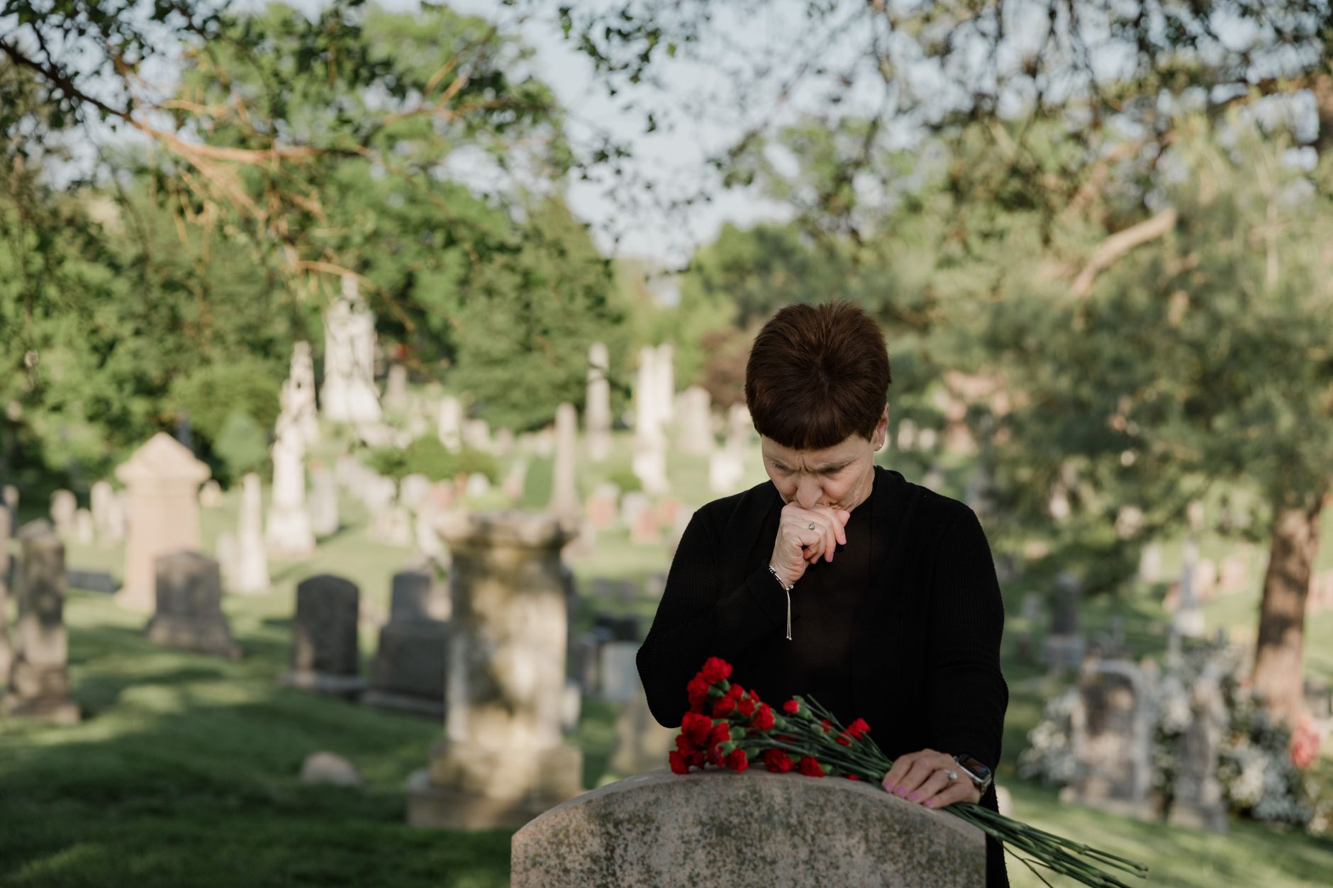 A woman in black paying her respects at a grave, holding flowers in a serene cemetery.