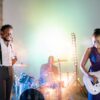A vibrant live band performance featuring a singer, guitarist, and drummer on stage with colorful lighting.