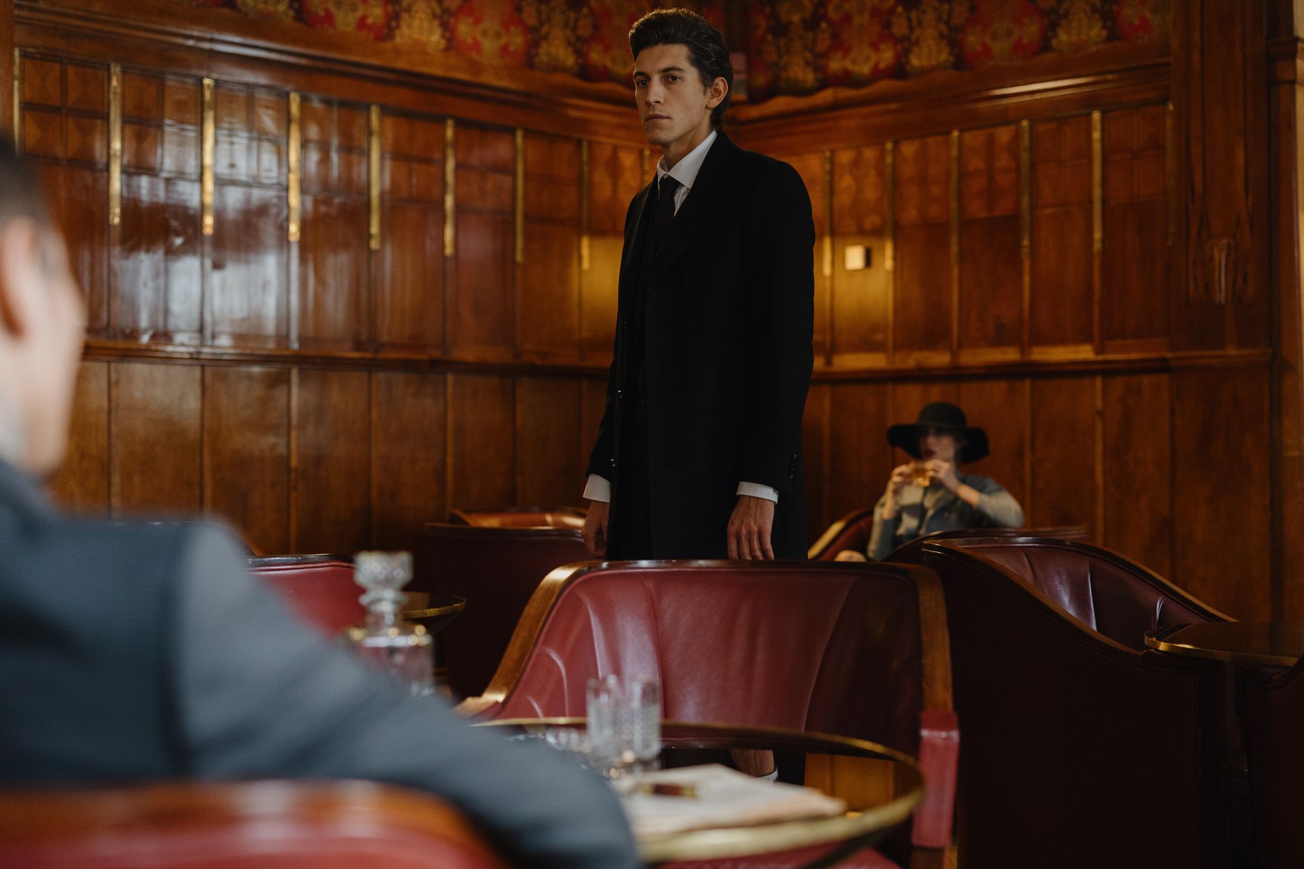 A man in a suit stands confidently in a vintage-style lounge with wooden panels and elegant decor.
